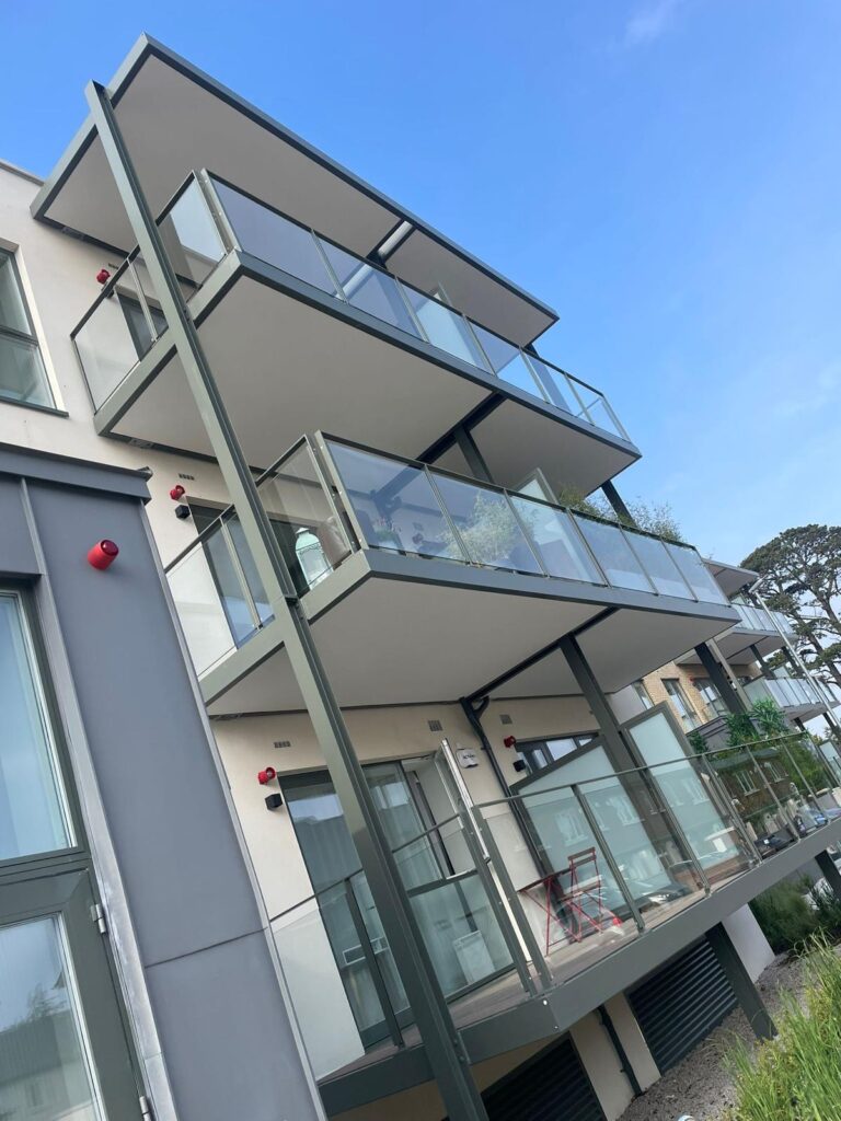 A white building with three glass balconies on it.The balconies have dark grey details on them.A long dark-grey beam connects them all.There is a dark grey room at the very front of the building.