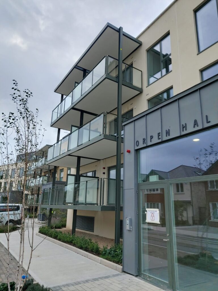 There is a dark grey room at the front.It has the words 'ORPEN HALL' on it.It has a glass door on the front of it.The bbuilding connected to the grey doorway is a sandy colour.It's three stories tal with balconies with glass and dark grey balconies.