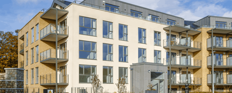 A photo of a building. One wall is white and the other two are sandy coloured bricks.There is balconies with glass banisters.There is a dark grey wall on the roof with glass railings in front of it.There is a dark grey room at the front.It has the words 'ORPEN HALL' on it.