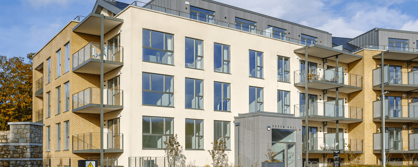 A photo of a building. One wall is white and the other two are sandy coloured bricks.There is balconies with glass banisters.There is a dark grey wall on the roof with glass railings in front of it.There is a dark grey room at the front.It has the words 'ORPEN HALL' on it.