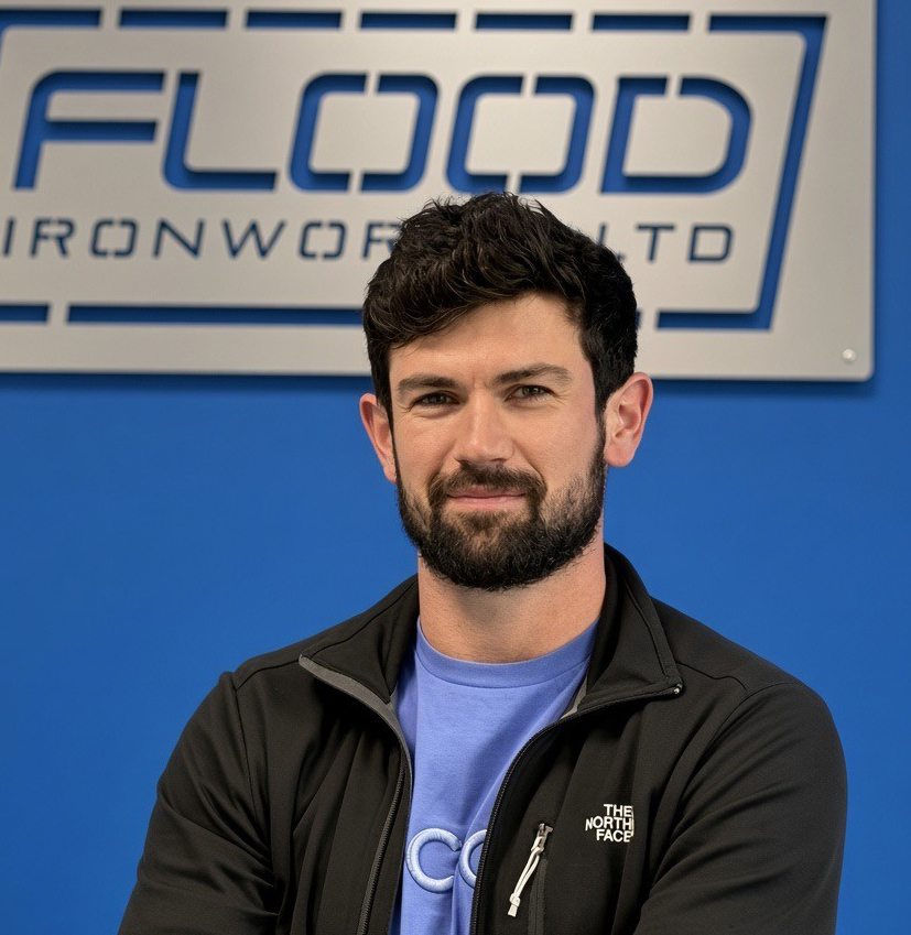 man with black hair and beard weaing a black jacket and a blue top