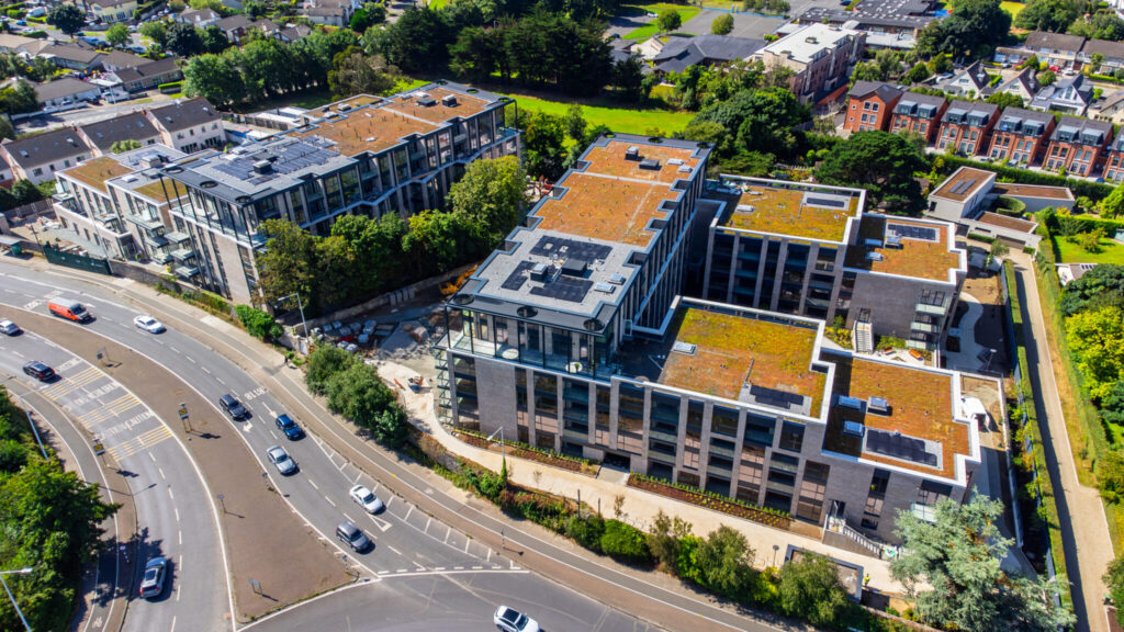 A photo of a building.It is two U-shaped buildings facing each other.They have solar panels on the roof.The height of the building gradually decreas as it reaches either end.They have windows a light grey brick walls.There is a road on the left side of the photo.Ther is houses in the background.