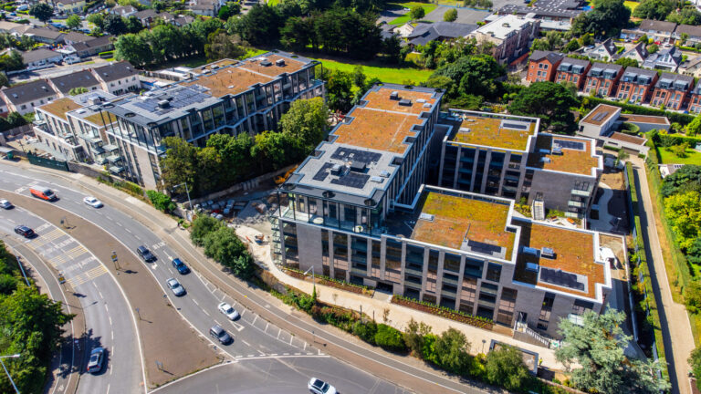 A photo of a building.It is two U-shaped buildings facing each other.They have solar panels on the roof.The height of the building gradually decreas as it reaches either end.They have windows a light grey brick walls.There is a road on the left side of the photo.Ther is houses in the background.