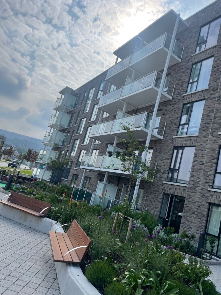 A picture of apartment blocks.They have modern grey brick work.They have white balconies with privacy screens on either side and glass banisters on the front.There is greenery and plants in front of the building.There is wooden benches on the wall in front of the plants.