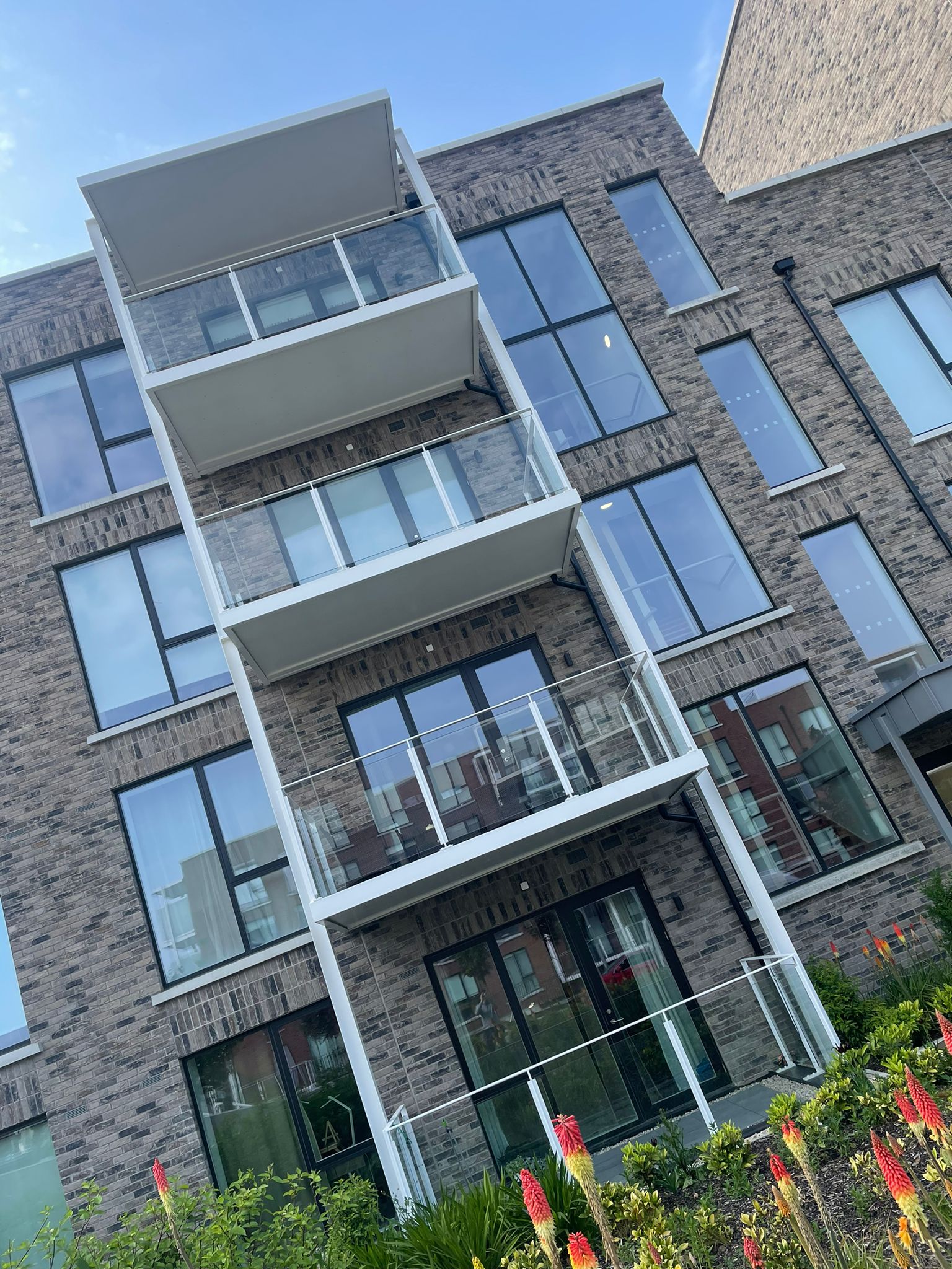 A slanted photo of apartments.They have balconies with glass banisters.They have dark grey brick work.There is plants in front of the building.