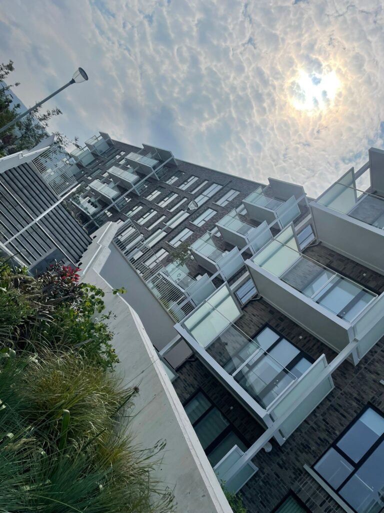 A slanted photo of apartment blocks.They are moder, grey bricked.They have white balconies.There are green plants in front of a tall white wall.On the left of the wall there is stairs in the left middleground.