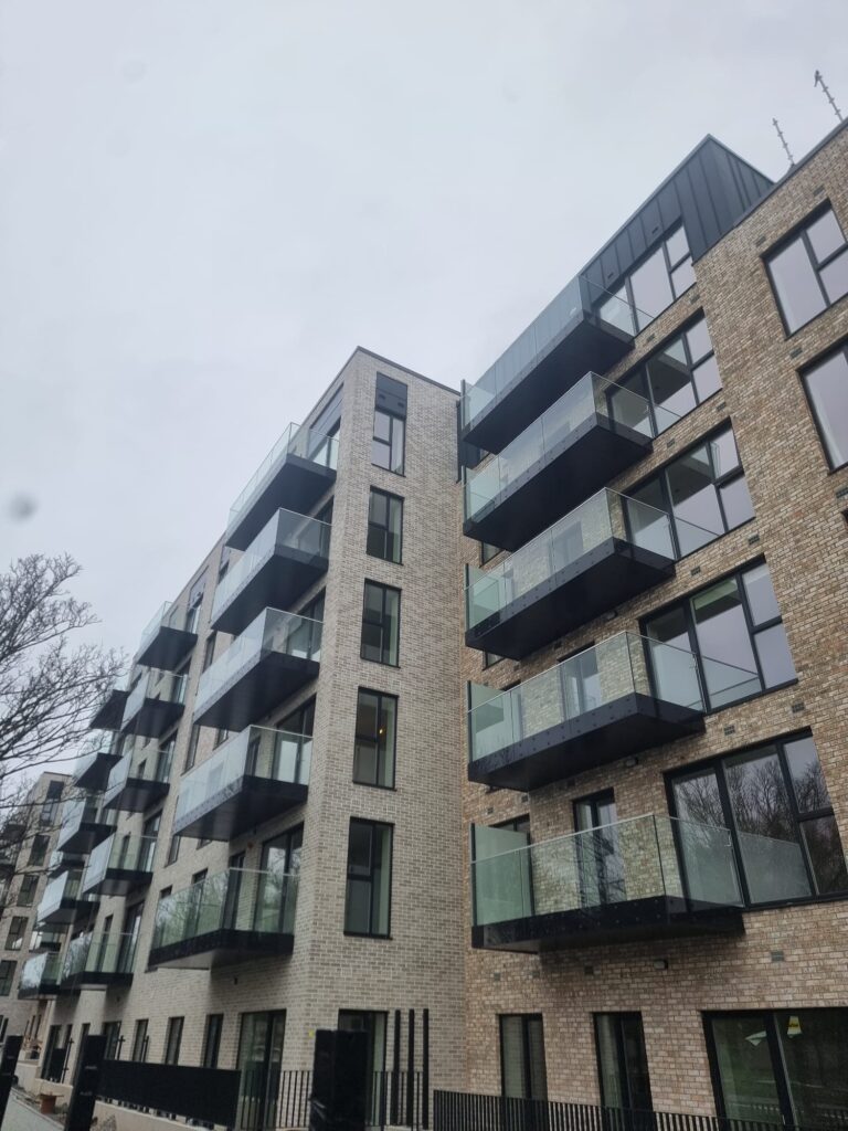 A photo of apartment buildings.They have a sandy brick colour.They have balconies with glass banisters. They have big windows on the building.The building is 6 stroies tall.