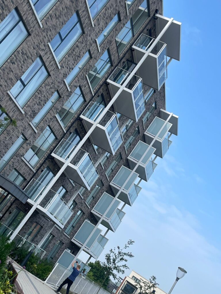 A picture of apartment blocks.They have modern grey brick work.They have white balconies with privacy screens on either side and glass banisters on the front.A man at the bottom is taking a photo of the buildings.