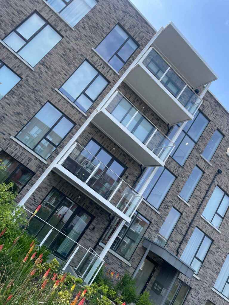 A slanted photo of apartment blocks.A picture of apartment blocks.They have modern grey brick work.They have white balconies with privacy screens on either side and glass banisters on the front.There is green plants in front of the buildingsThe entrance tothe building is on the right.