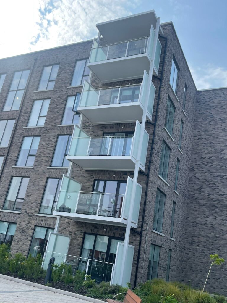A picture of apartment blocks.They have modern grey brick work.They have white balconies with privacy screens on either side and glass banisters on the front.There is greenery and plants in front of the building.