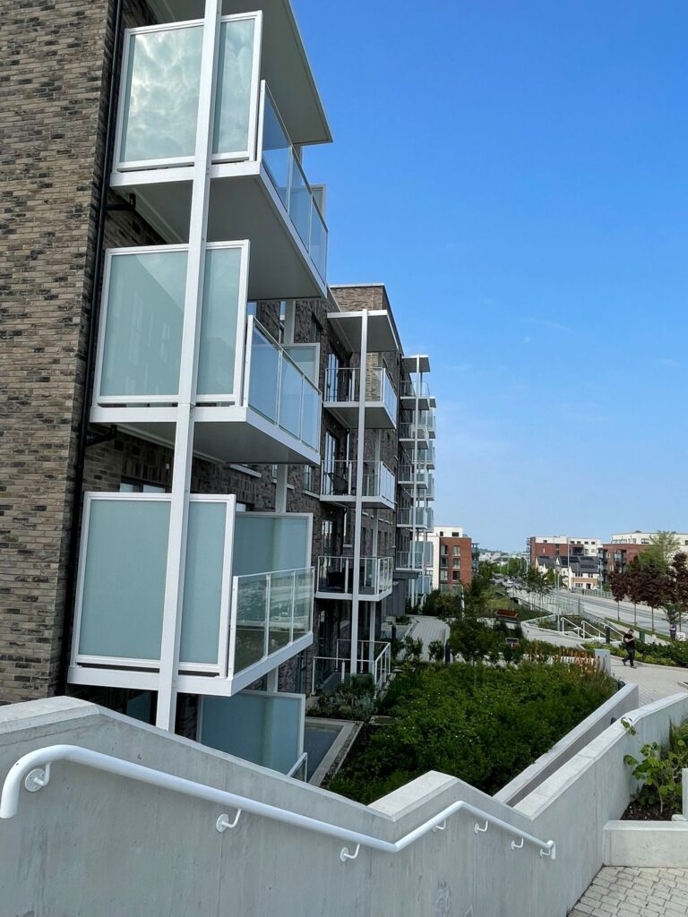 A picture of apartment blocks.They have modern grey brick work.They have white balconies with privacy screens on either side and glass banisters on the front.There is a walled area with greenery and plants.There is a big staircase with white railings and white brick work.