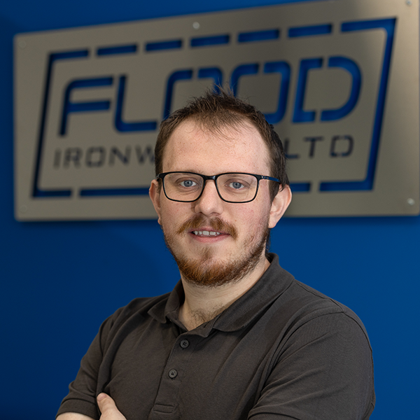 A man in a grey-brown shirt.He has brown hair and a ginger beard.He wears glasses.