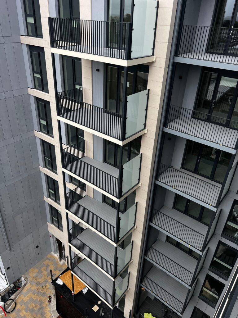 A tall modern apartment building. Each floor has a small balcony with black railings and large glass doors .The middle section is slightly out further than the rest.The photo is taken at a higher angle facing downwards over looking the apartment block