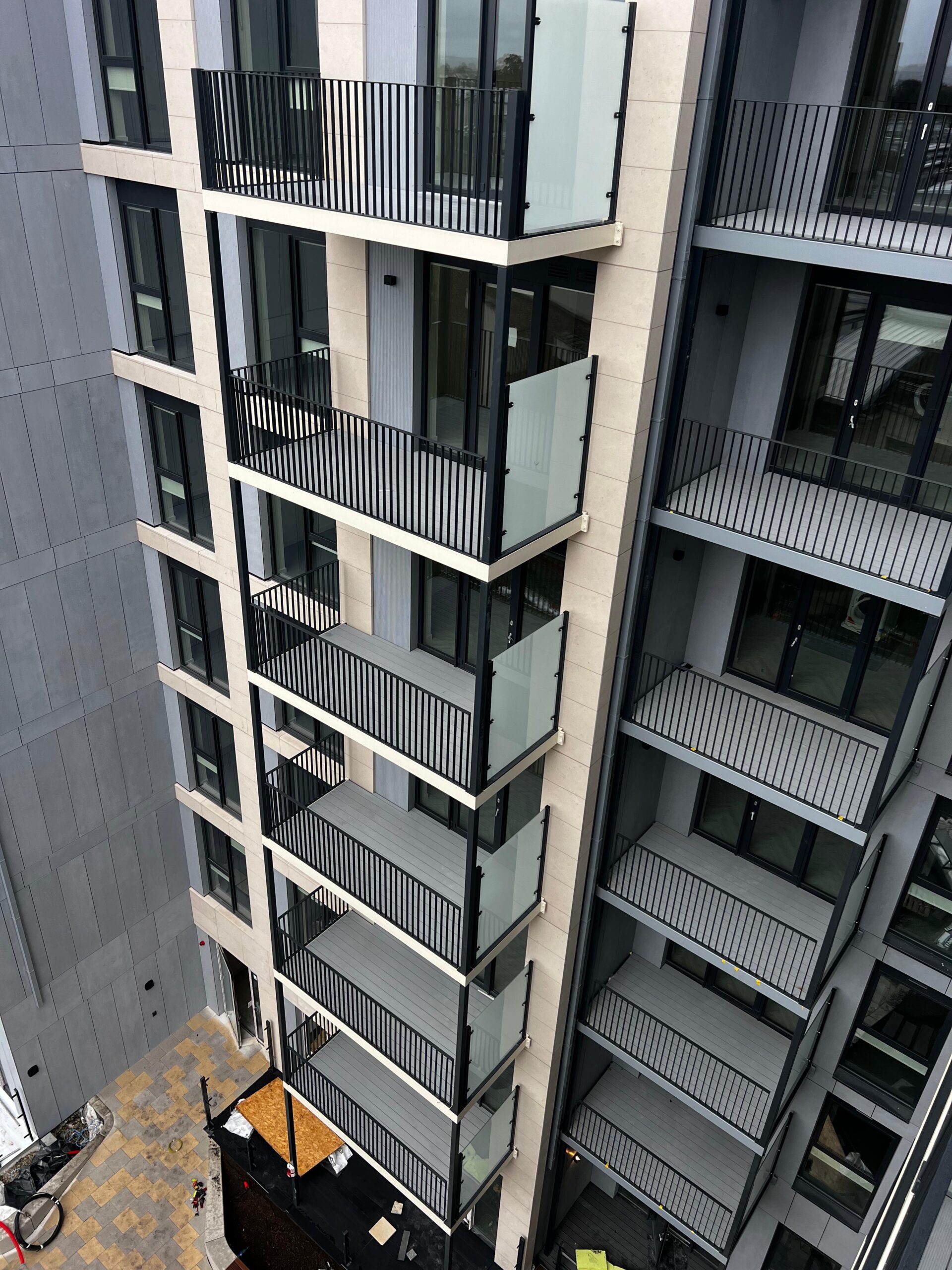 A tall modern apartment building. Each floor has a small balcony with black railings and large glass doors .The middle section is slightly out further than the rest.The photo is taken at a higher angle facing downwards over looking the apartment block