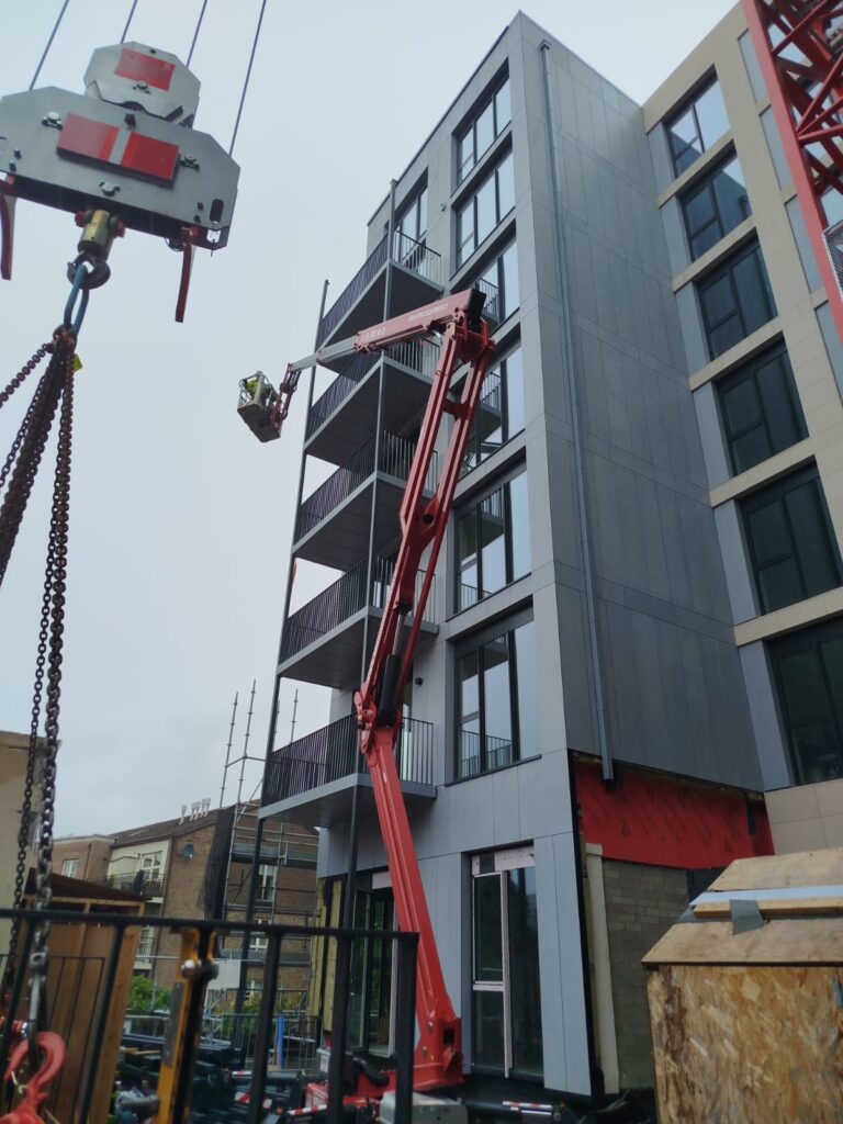 A tall building with a line of balconies one above the other.The balconies have dark grey railings.The building is light grey.There is a red scissor lift going up along the building.A hook of a crane is visible on the floor.
