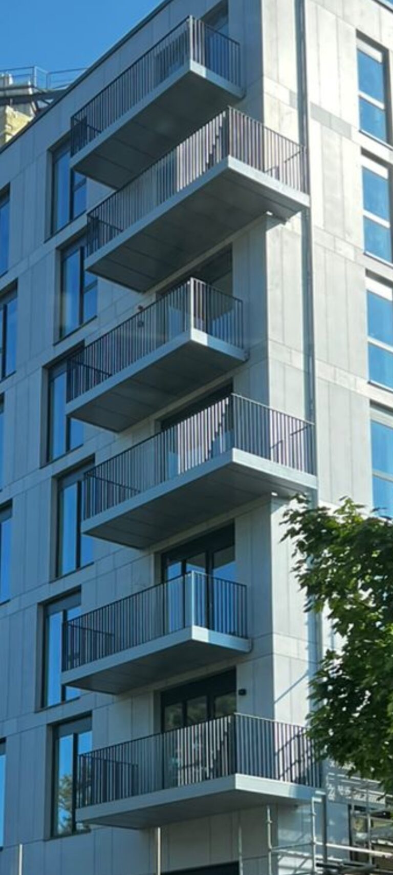 A tall building with a line of balconies oneabove the other.Every second balcony is larger and wraps partially around the building.The balconies have dark grey railings.The building is light grey.