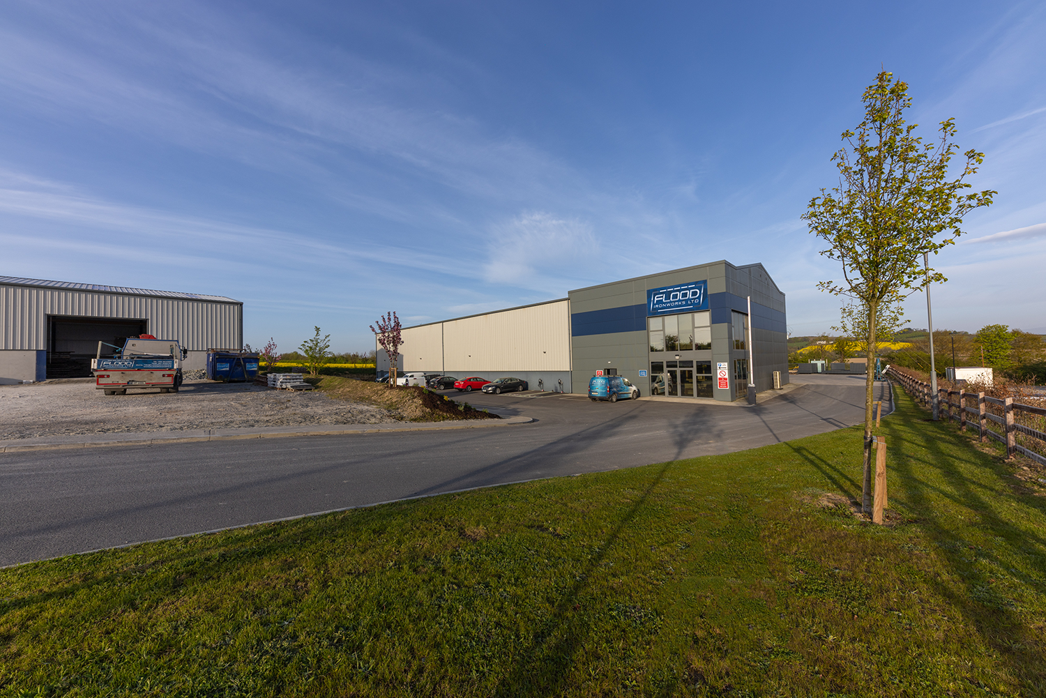 A grey building with a corragated metal garage attached to it.There is a blue stripe on it with a blue sign saying 'FLOOD IRONWORKS LTD'There is a white line below it and a white dashed line above it, connected by two grey lines.There is a small line of cars parked in front of the building.There is a corragated metal garage with a truck outside of it.There is a patch of grass with a small tree where the photo is taken from.