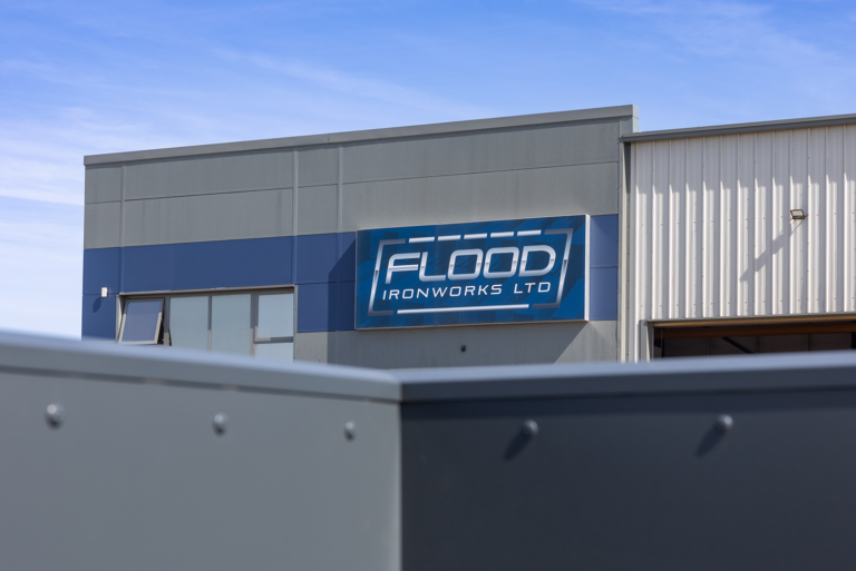A grey building with a corragated metal garage attached to it.There is a blue stripe on it with a blue sign saying 'FLOOD IRONWORKS LTD'There is a white line below it and a white dashed line above it, connected by two grey lines.There is a dark grey structure blocking the bottom half of the cameras view.
