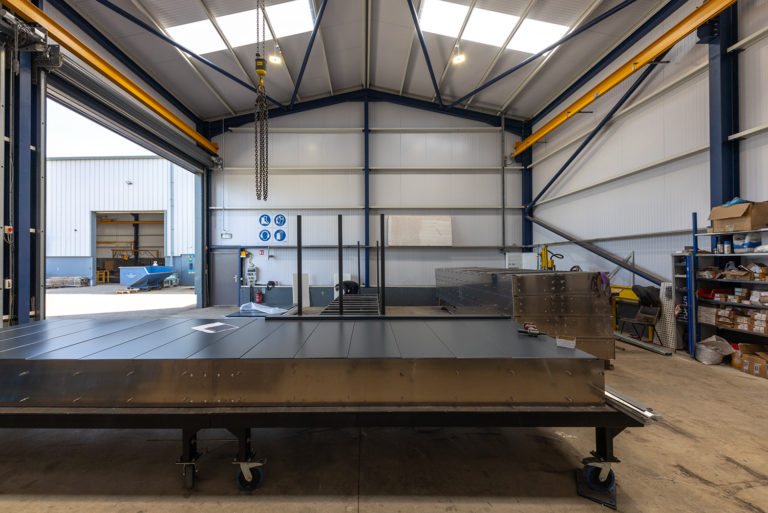 A large iron sheet on a wheelie trolly.It is in a large garage with grey walls.One garage door is open,showing anither similar grey building.A man is working in the background.There is shelves with boxes on them to he right.More large iron sheets are on the right.