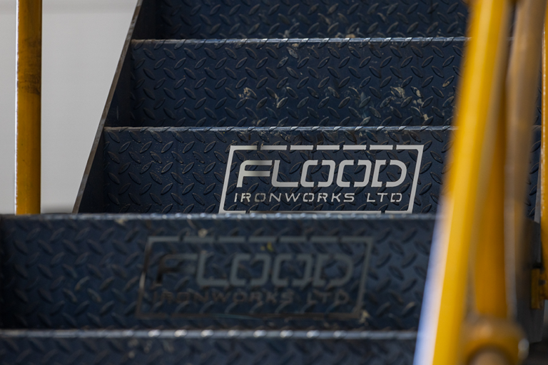 A close up of a blue staircase.The words 'FLOOD IRONWORKS LTD' are on the stairs.There is a yellow railing on either side of the stairs.The stairs are metal and have a leaf shaped design on them.