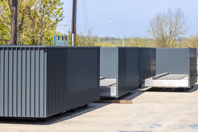 A group of grey containers outside on a concrete floor.They have a light grey ,iron floor.There is trees in the background.