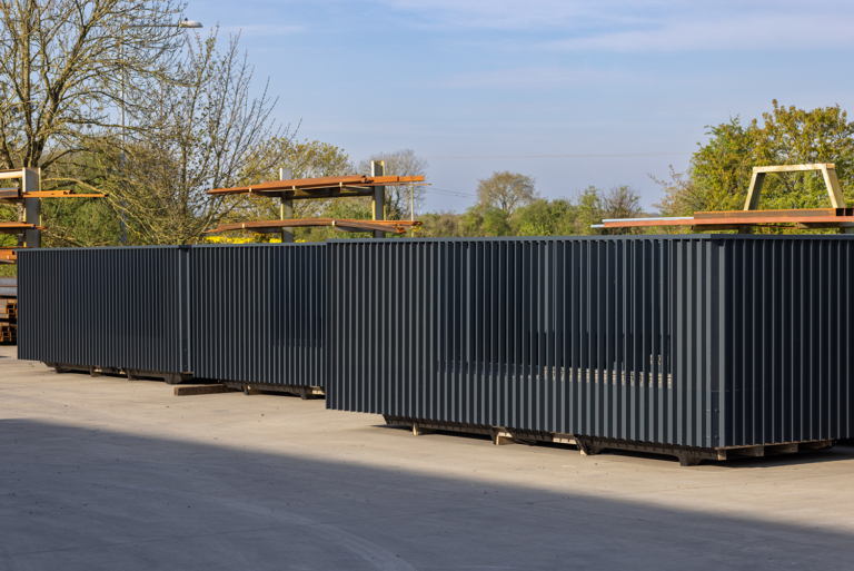 A group of dark grey metal structures.They have a fence style pattern with gaps between each pillar.There is wooden frames behind the metal structures.