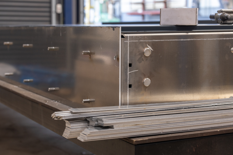 A close up shot of the corner of a metal structure. There is bolts coming out of the metal sheet on the left side of the image. There is small metal strips in front of the metal structure.