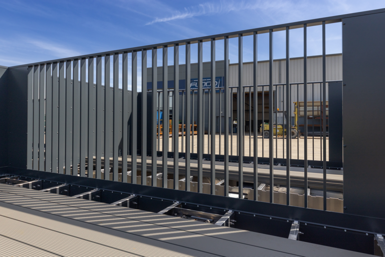 A large,dark grey metal structure.It has a fence-like pattern in the middle.In the back ground there is a Flood Ironworks building.It's grey and dark grey.There is a yellow forklift in front of the building.In front of the metal structure there is a wooden patio floor.