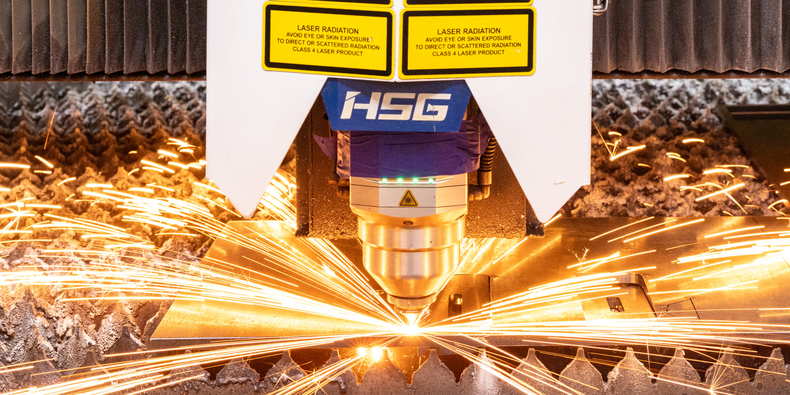 A close-up of an HSG laser cutting machine cutting metal, with bright orange sparks flying outward around the cutting head.