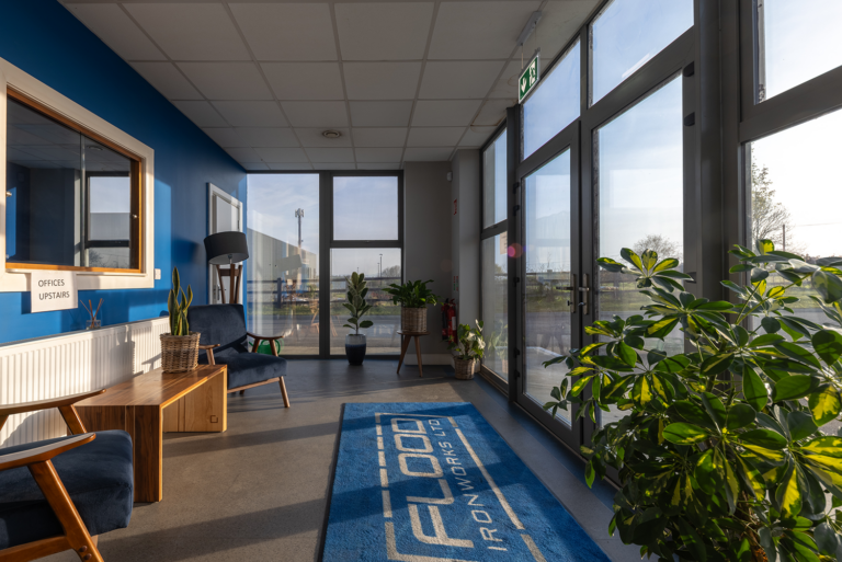 A doorway with glass and a dark grey frame.There is a rug on the floor whick is blue and has the words 'FLOOD IRONWORKS LTD' on it.There is two gey-blue seats and a wooden desk.There is a mirror with a sign below saying 'Office Upstairs.'
