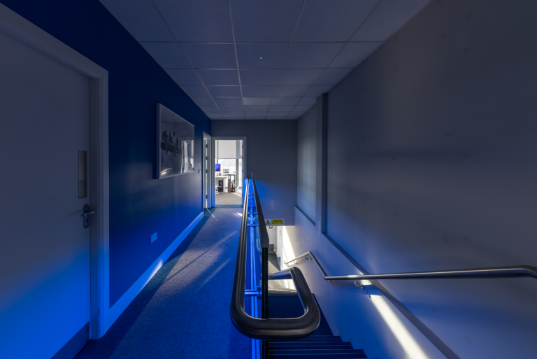 A staircase and hallway.There is a royal blue light shining on the stairs and floor.The carpet on the floor is grey and the stairs is dark grey.There is a hand rail on both sides of the stairs.There is a a closed white door on he left.At the end of the hallway there is an open door leading to an office with a desk and computer.The walls are dark grey with white trimmings.