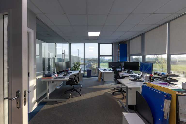 A picutre of and office with three desks and computer set ups.There is windows on every wall.There is a blue storage unit in the back right of the room.Eaxh desk is white and the floor is a grey carpet.The white door to the room is open and there is a plant inthe left corner.