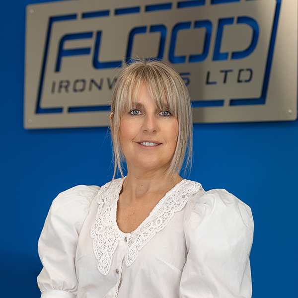 A woman with blonde hair and a fringe.She is wearing a white blouse.She is looking at the camera with her arms by her side.