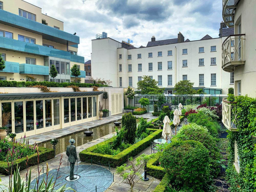 A garden with a statue on a circular,dark grey floor.There is railings around the statue. There is green bushes in rectangular patterns with plants in the middle.There is a paved patio with a pond with fountains in it.There is a white building in the back with old fashioned windows.To the left there is a yellow building with balconies.The balconies have blue glass railings.There is a pale yellow building with seats and tables looking out at the garden.