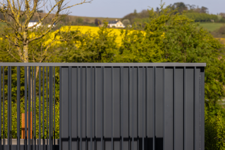 A photo of a dark-grey metal structure.One side has closed ridges on it.The other side has an open gap design.There is trees in the background.