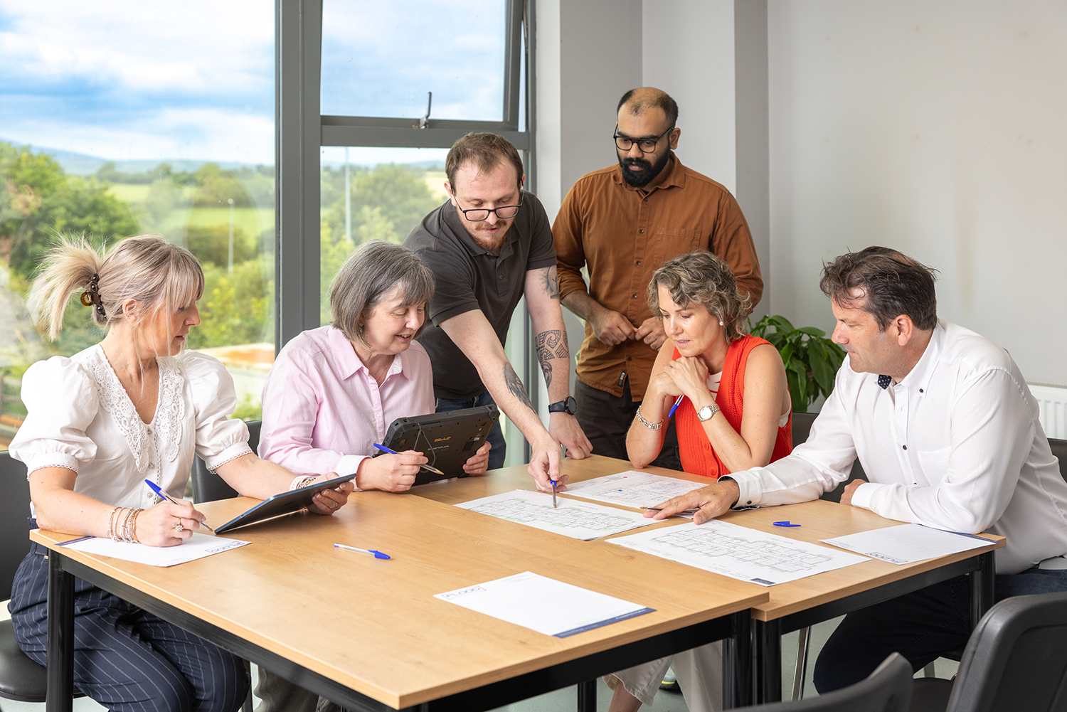 A group of 6 people around a table working together.The woman on the left has blonde hair in a clip.She is wearing a white top and navy stripey pants.She is holding an ipad and is writing on paper with a pen.The woman next to her has short grey hair in a bob.She has a pink shirt on.She is holding an Ipad.Theman in the middle is standing up and pointing at a page.He has a grey shirt on.He has brown hair,a beard and glasses.He has tattoos on his arms.There is another mane behind him.He has a brown shirt and glasses on.He is bald and has a black beard.The woman next to him has short brown hair.She has an orange top on.The man next to her has a white shirt on and brown hair.