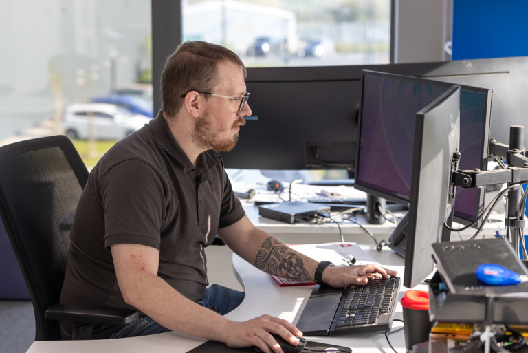 A man working at a computer setup.He has brown hair and a beard.The man is in a brown collared shirt.He has glasses on.He has a watch on his left arm and a tattoo.