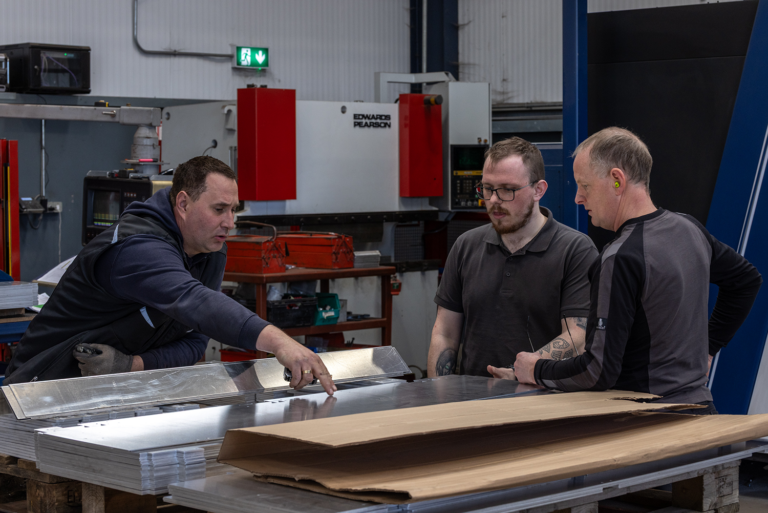 Three men working on a piece of metal together.One is wearing ear protections,another is wearing glasses and the other is pointing at the sheet of metal.