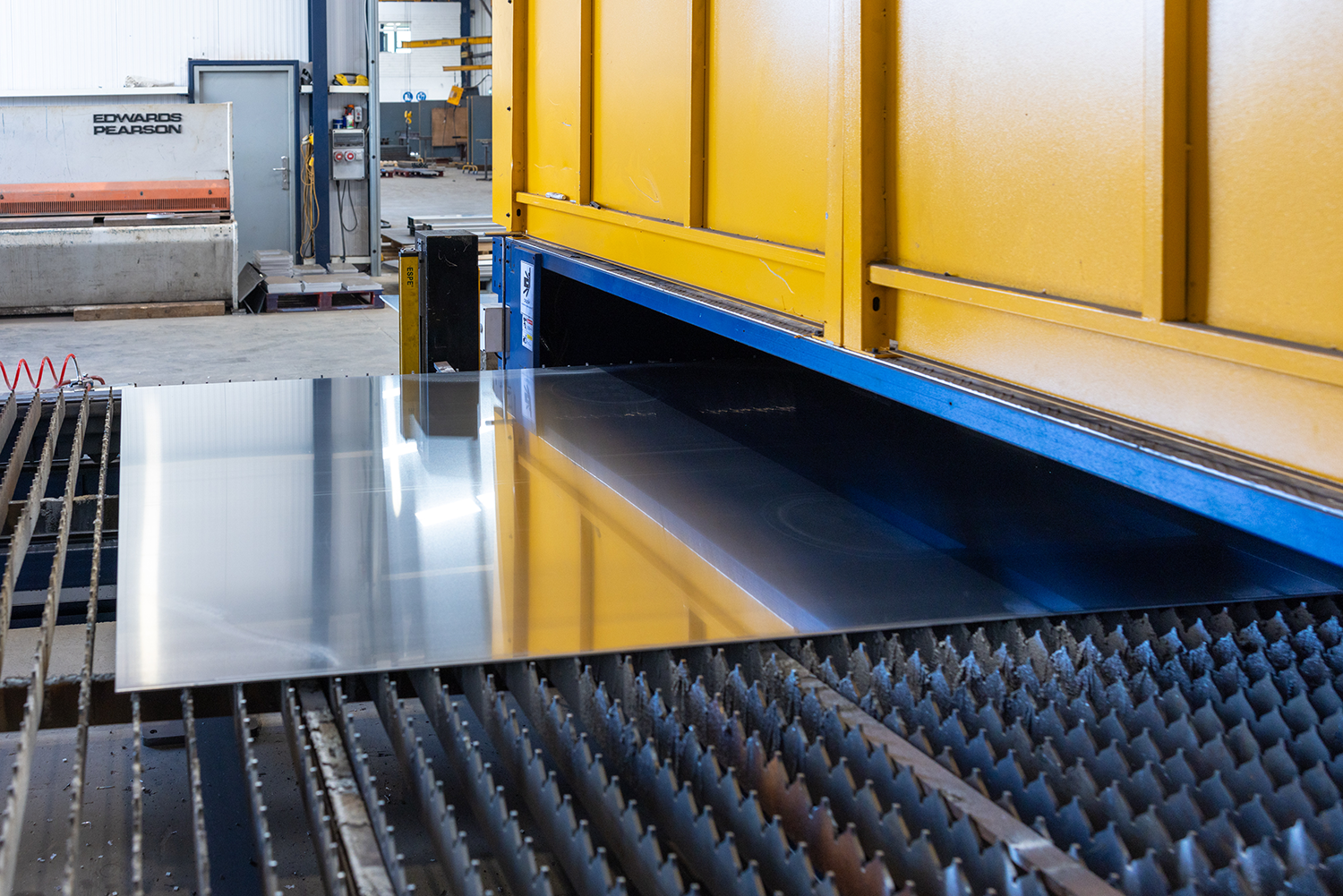 A sheet of metal on a conveyor belt going underneath a yellow machine.