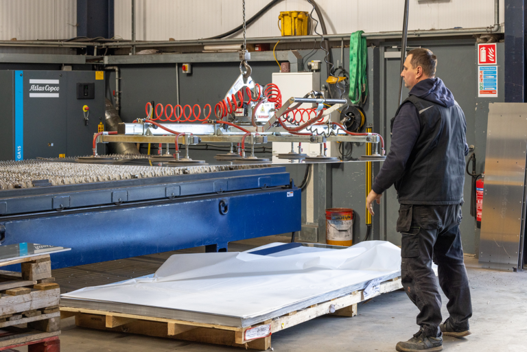 A worker stands beside an industrial machine with a vacuum lifting system, handling large sheets of metal in a manufacturing workshop.