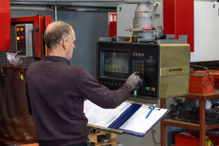 A worker wearing ear protection operates an industrial control panel beside a manufacturing machine, with technical drawings and a pen on the workstation.