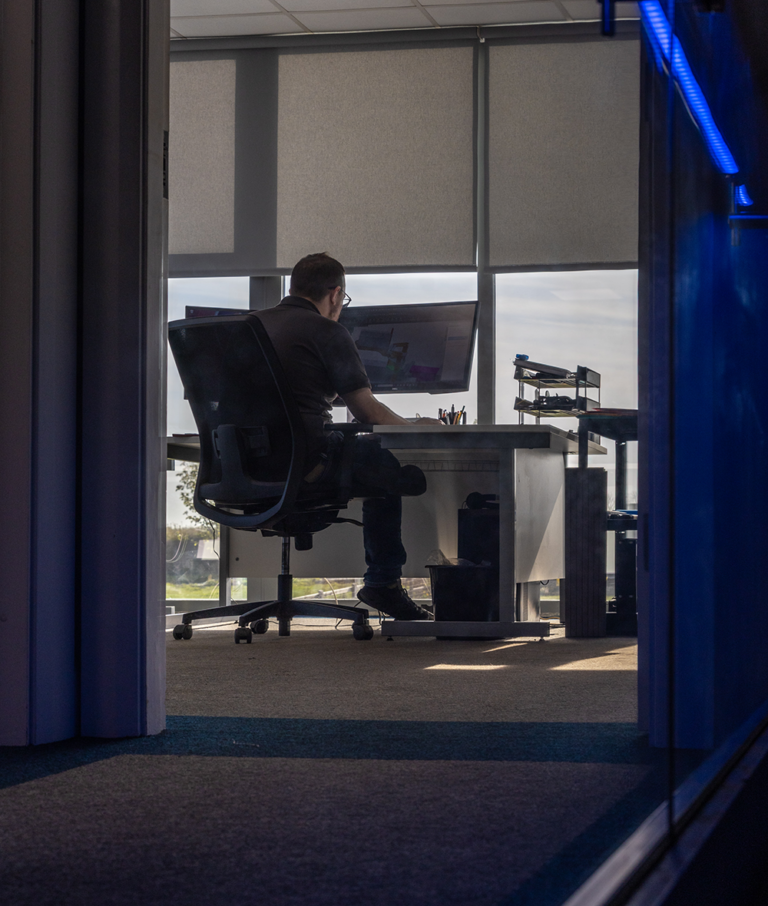 A man working on a computer.He is wearing a grey shirt and sitting on a black wheelie chair.He has brown hair and is wearing glasses.The photo is taken from behind the man.