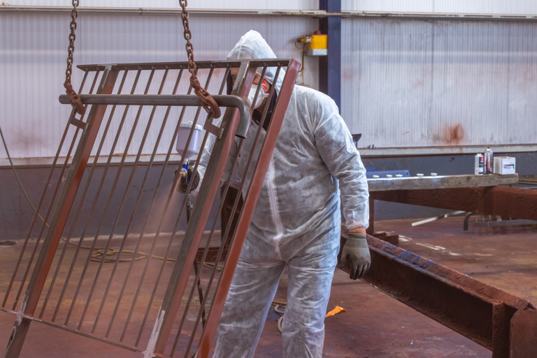 A man in a white protective suit from head to toe is spray painting a fence brown in a large shed.He is wearing gloves and a mask.