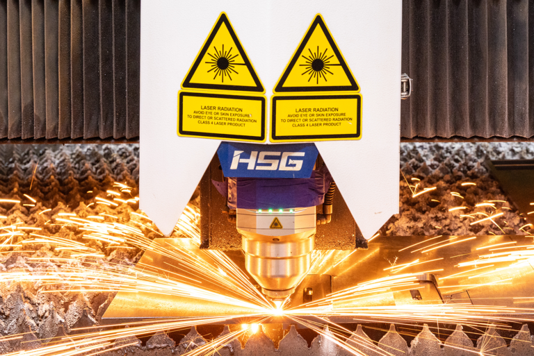 A close-up of an HSG laser cutting machine cutting metal, with bright orange sparks flying outward around the cutting head.