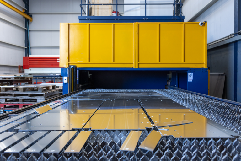A sheet of metal on a conveyor belt going underneath a yellow machine.