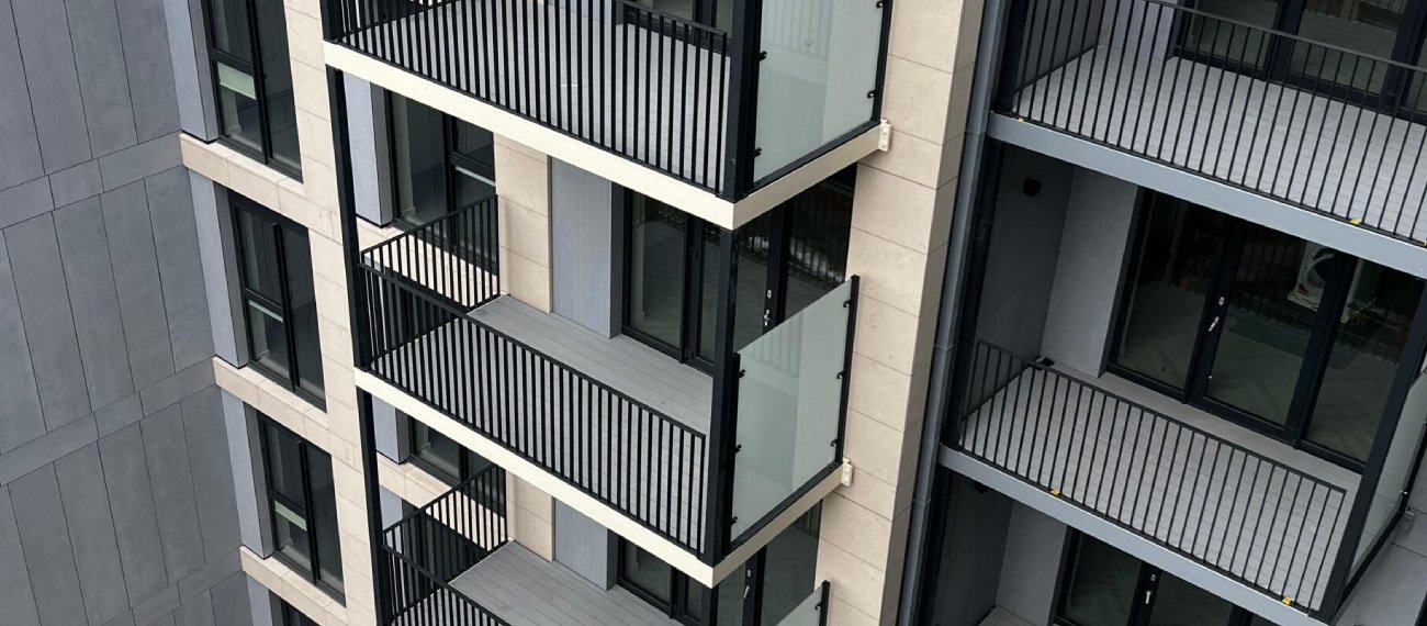 A tall modern apartment building with several floors. Each floor has a small balcony with black railings and a big glass door. the building is mostly grey and white. The middle block of the apartments is slightly more forward and sticking out . The photo is taken at a high angle facing down and over looking the apartment block