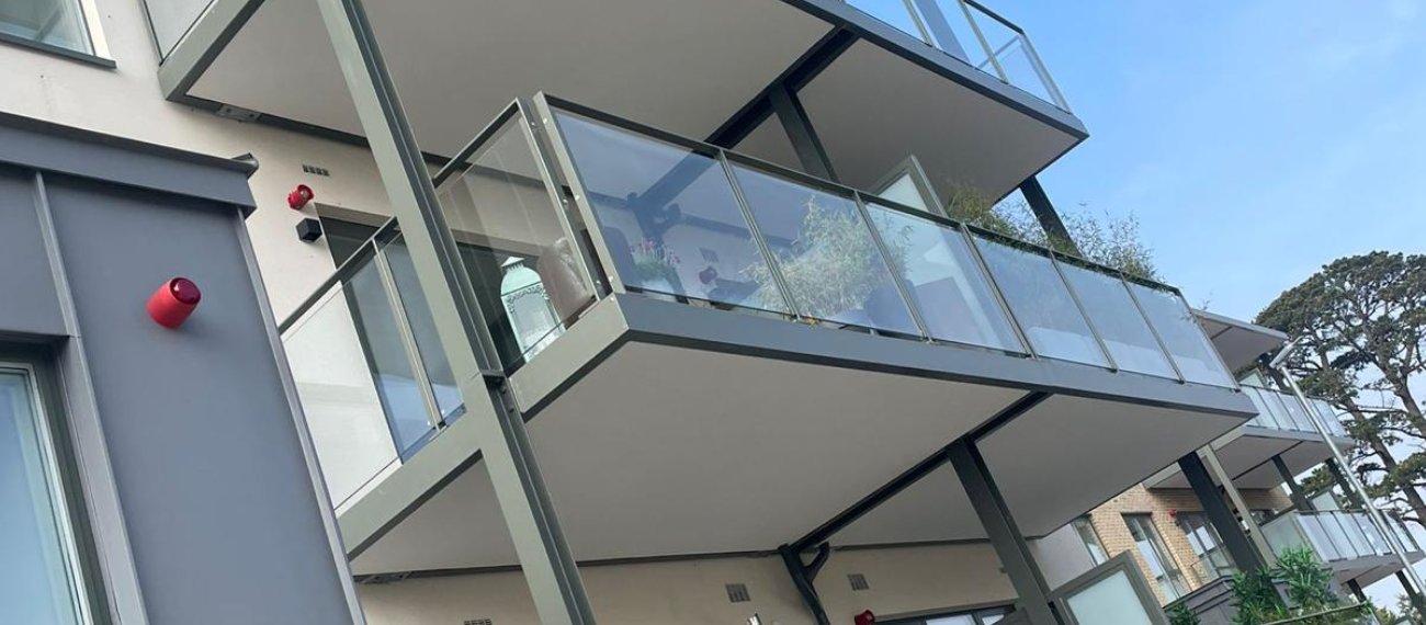 A zoomed in photo of an apartment balcony taken at a lower angle and facing upwards. The balcony has medium grey railings with a glass door and white wall