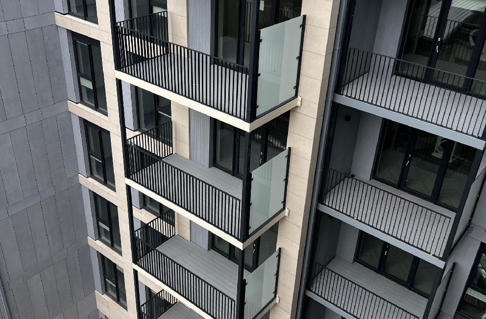 A tall, modern apartment building with several floors. Each floor has a small balcony with black railings and large glass doors. The building is mostly grey and white. The middle block of the apartments is slight more forward and sticking out. The photo is taken at a high angle facing down and overlooking the apartment block