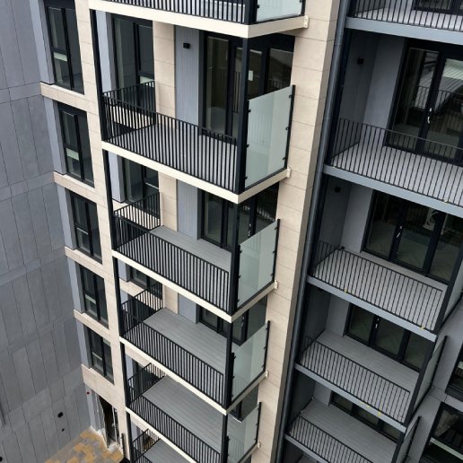 A tall modern apartment building with several floors . Each has a small balcony with black railings and large glass doors. The building is mostly grey and white .The middle block of apartments is slightly more forward and sticking out.The photo is taken at a a high angle facing down and overlooking the apartment block.